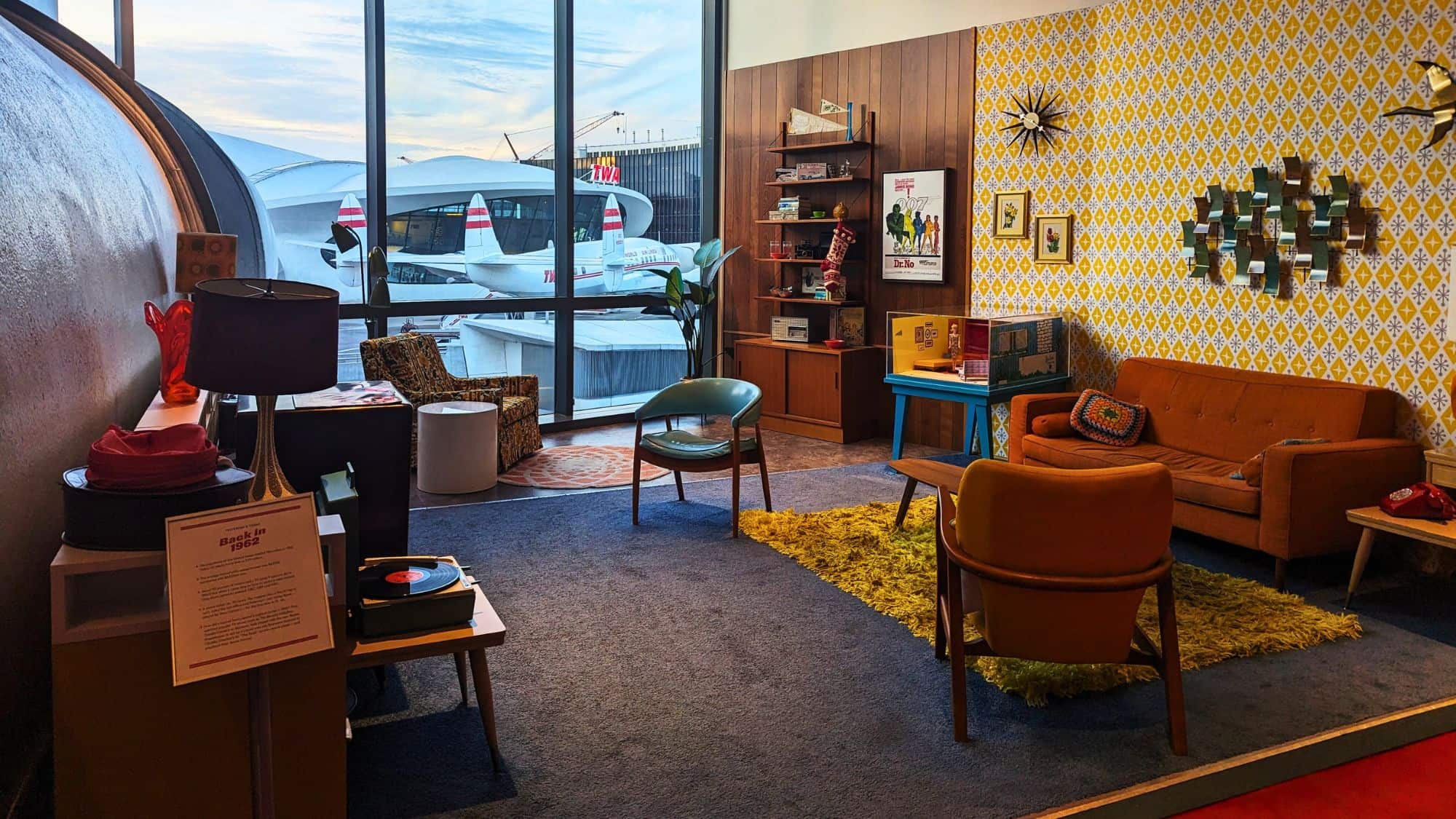 A 1960s-themed hotel room decorated with vintage furniture, mustard-colored shag carpet, a retro record player, and a wall of patterned wallpaper, with large windows showcasing a TWA airplane outside.