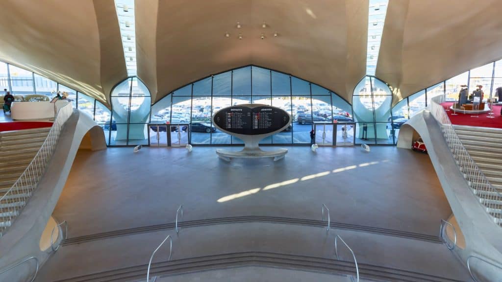 A spacious airport terminal with a sleek mid-century modern design, featuring a large split-flap departures and arrivals board in the center, surrounded by curved architecture and expansive glass windows.