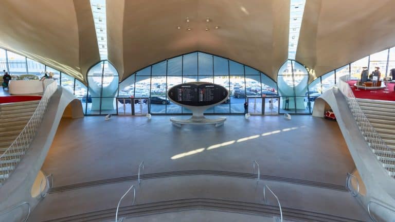 A spacious airport terminal with a sleek mid-century modern design, featuring a large split-flap departures and arrivals board in the center, surrounded by curved architecture and expansive glass windows.