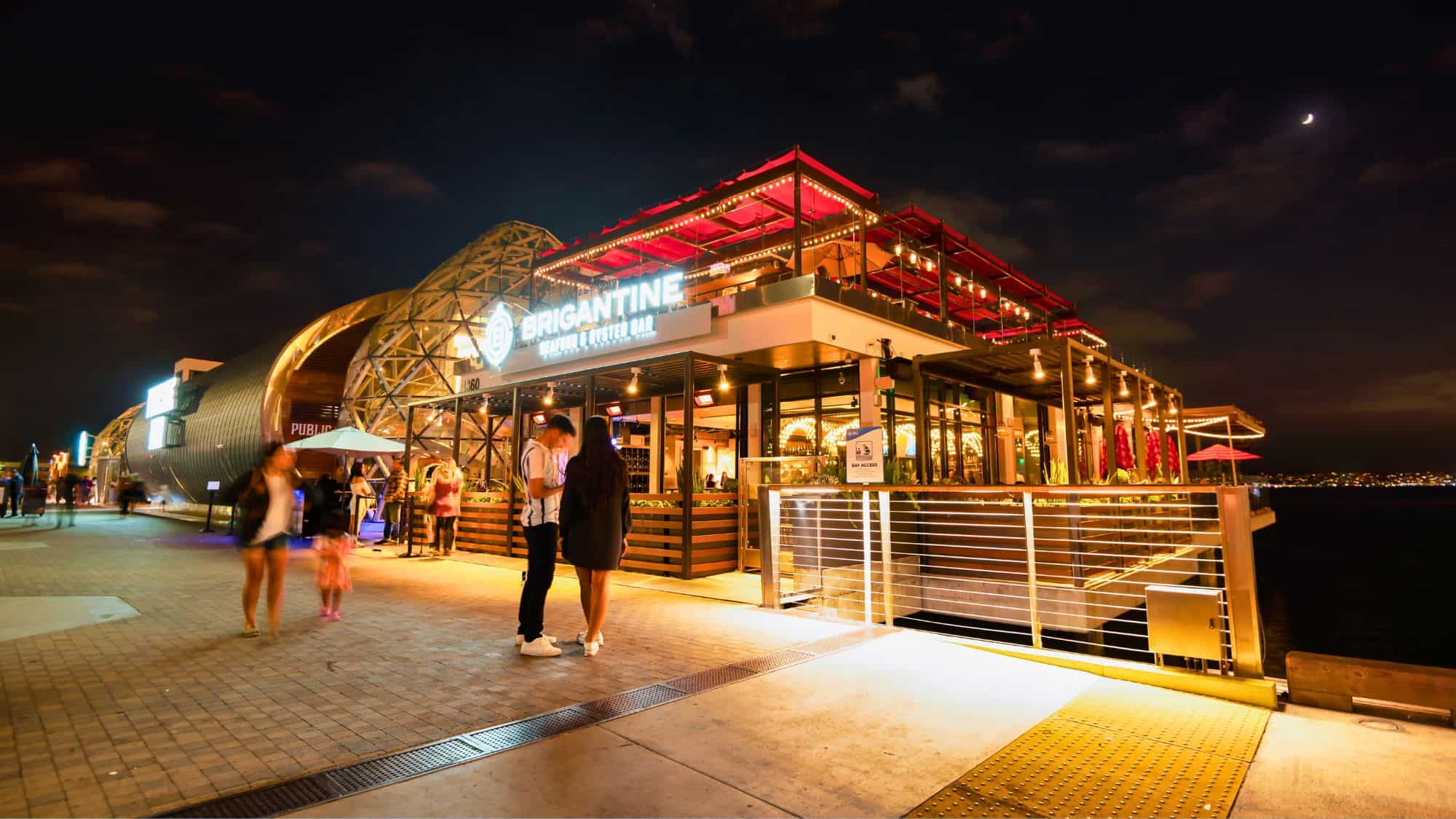 A modern seafood and oyster bar along the waterfront, lit up at night with a glowing sign reading "Brigantine," with people walking along the boardwalk under string lights.
