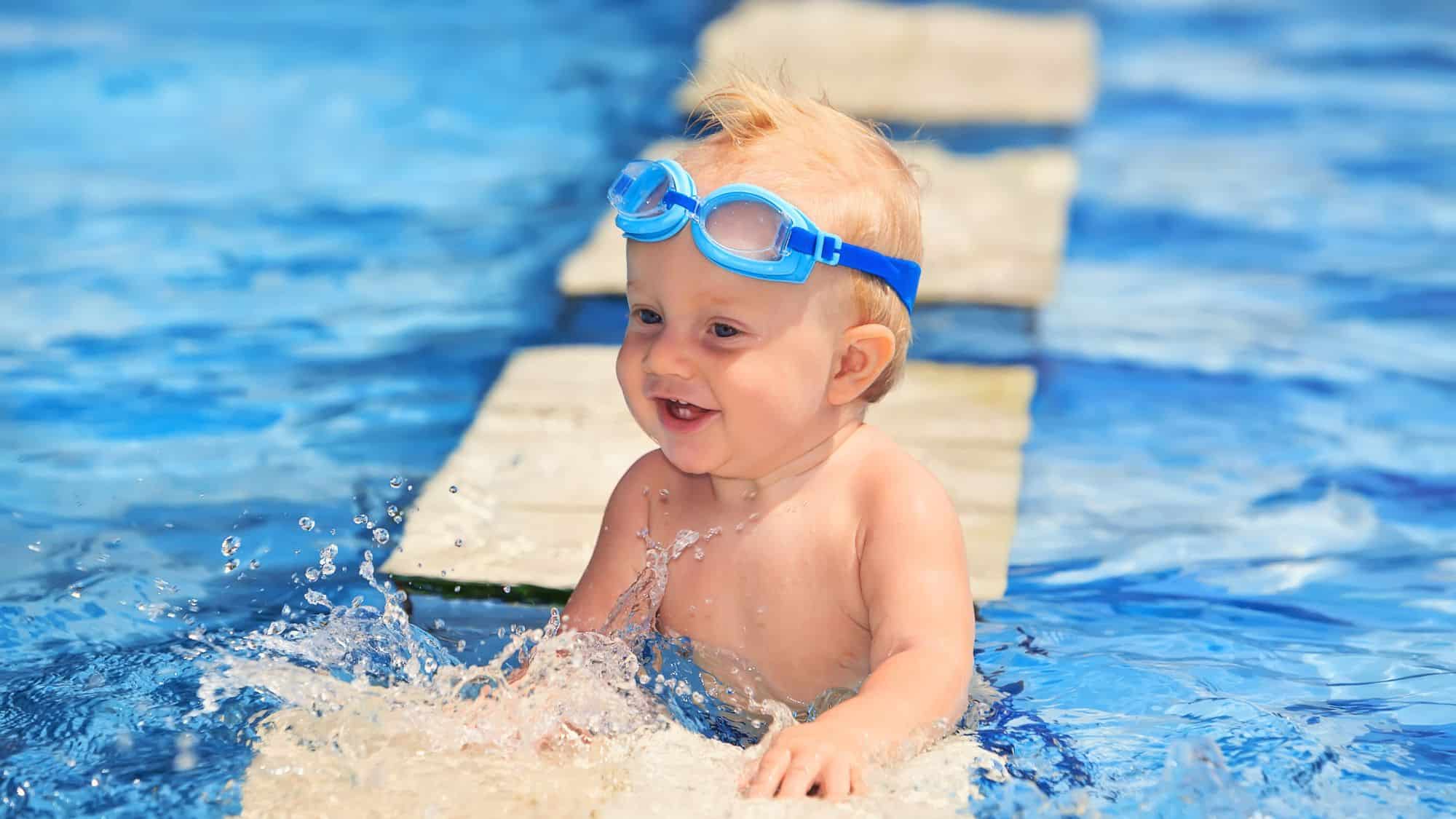 A happy toddler splashing in a pool while wearing blue goggles, grinning with excitement. The bright blue water and playful energy capture a fun swimming moment.