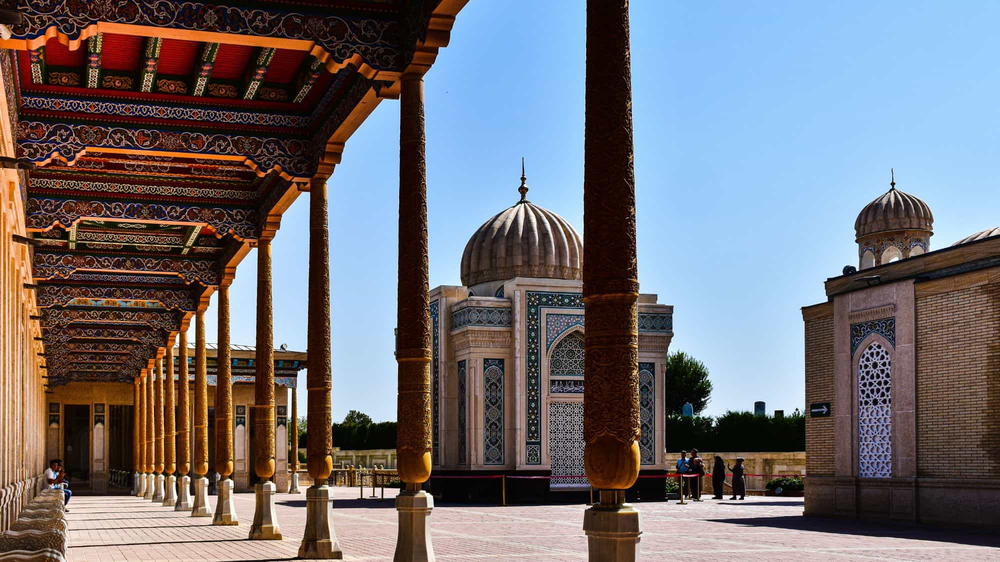 A beautifully detailed corridor leading to an ornate mausoleum in Uzbekistan, showcasing intricate Islamic architecture. The wooden columns and colorful tilework contrast with the clear blue sky.