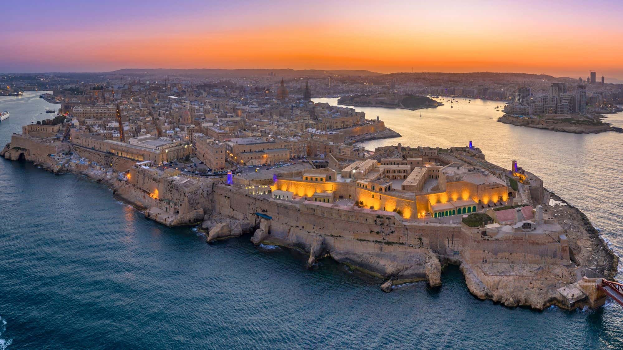 Sweeping aerial view of Valletta, Malta’s fortified capital, glowing under sunset skies with warm lights highlighting the city’s architecture and surrounding sea.