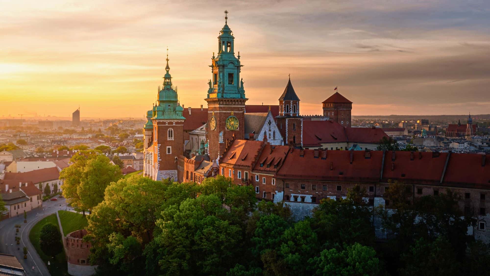 A panoramic view of Wawel Castle in Kraków, Poland, bathed in golden sunset light. The historic red-roofed castle towers over lush green trees, with the city skyline visible in the background.