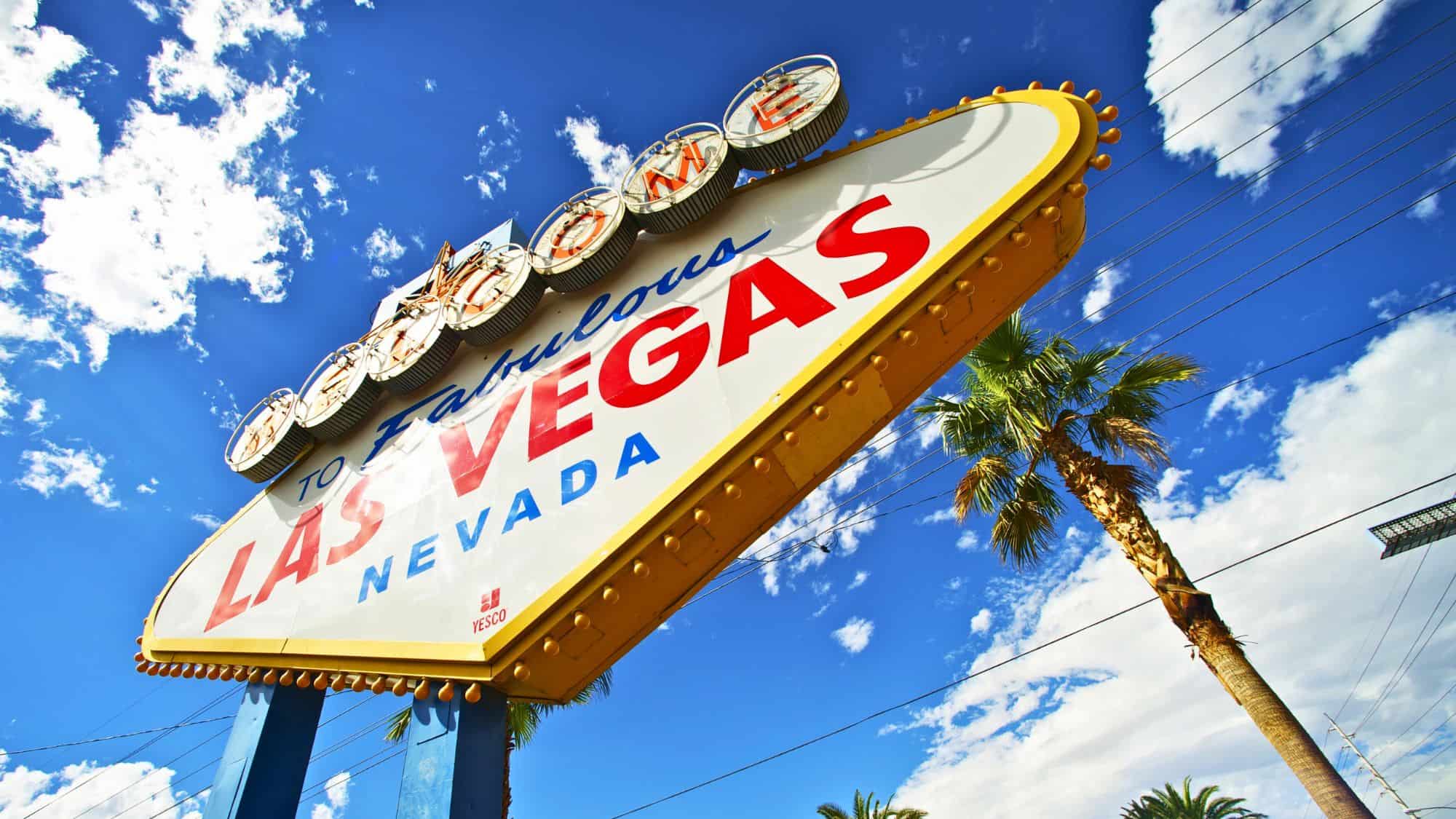 The famous "Welcome to Fabulous Las Vegas, Nevada" sign stands against a bright blue sky, framed by palm trees. The iconic landmark welcomes visitors to the entertainment capital of the world.