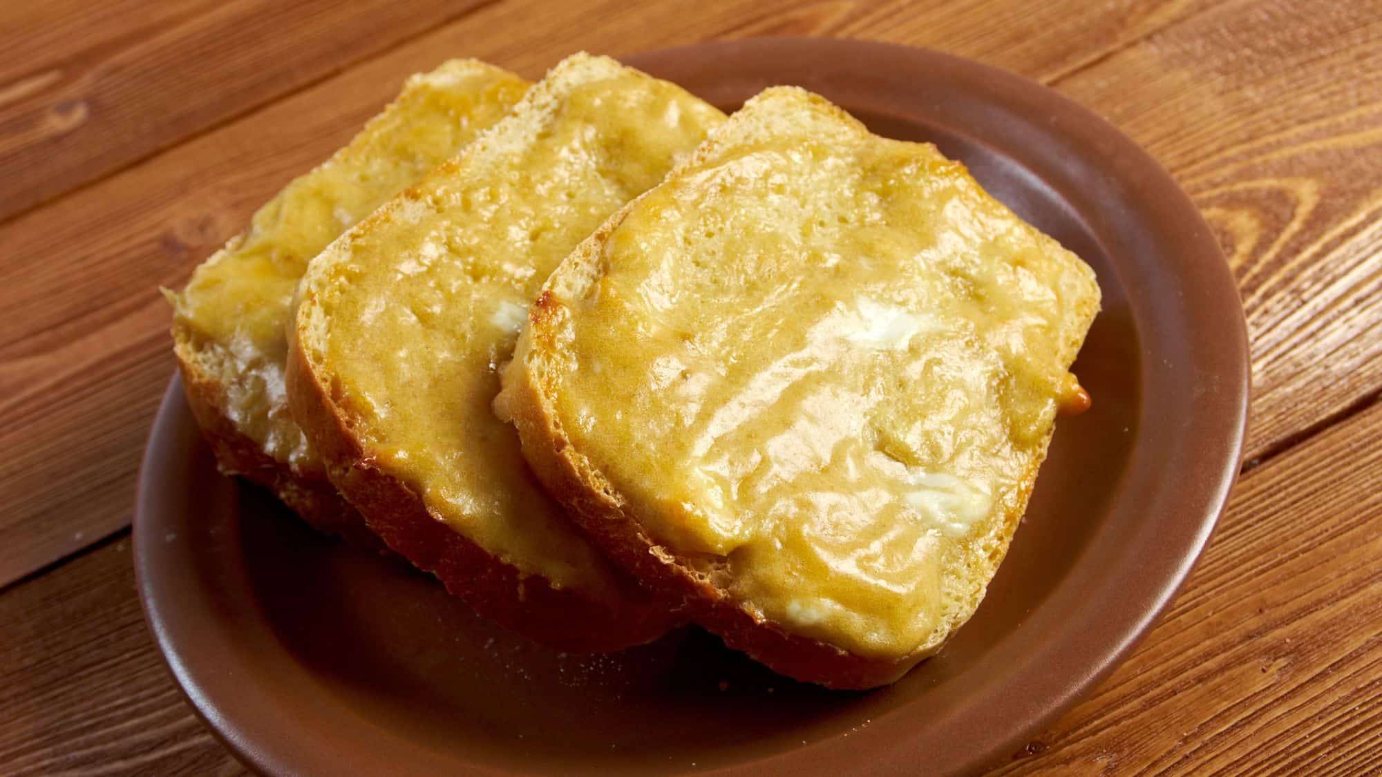 Three slices of toasted bread topped with a rich, golden cheese sauce, arranged on a brown ceramic plate atop a wooden table.