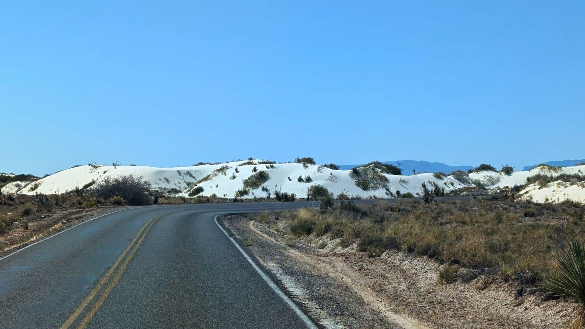 A winding road cuts through the desert landscape of White Sands National Park, bordered by bright white gypsum dunes dotted with shrubs under a clear blue sky.