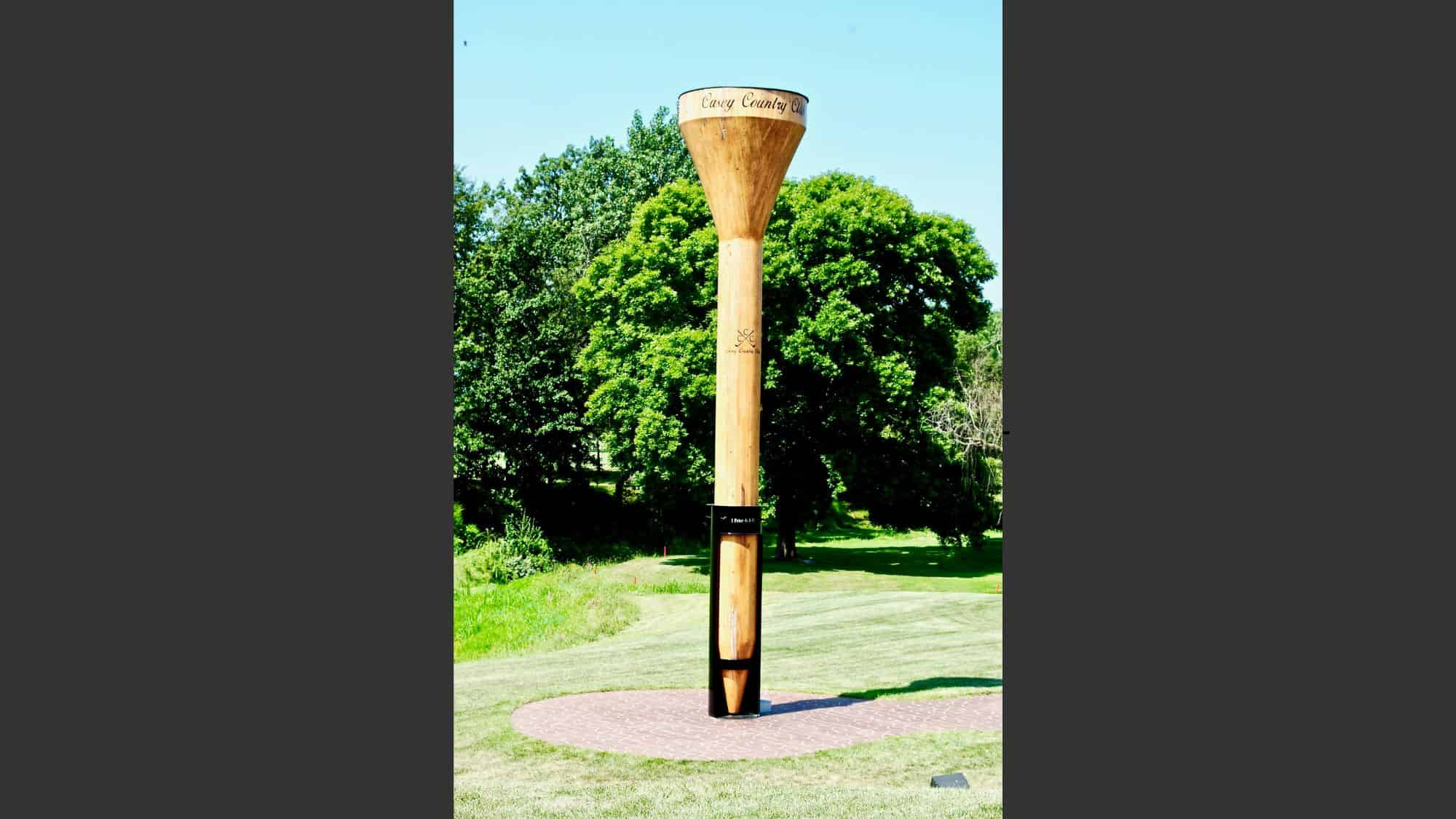 An oversized wooden golf tee sculpture stands upright on a circular brick platform in a green park setting, inscribed with “Casey Country Club.”