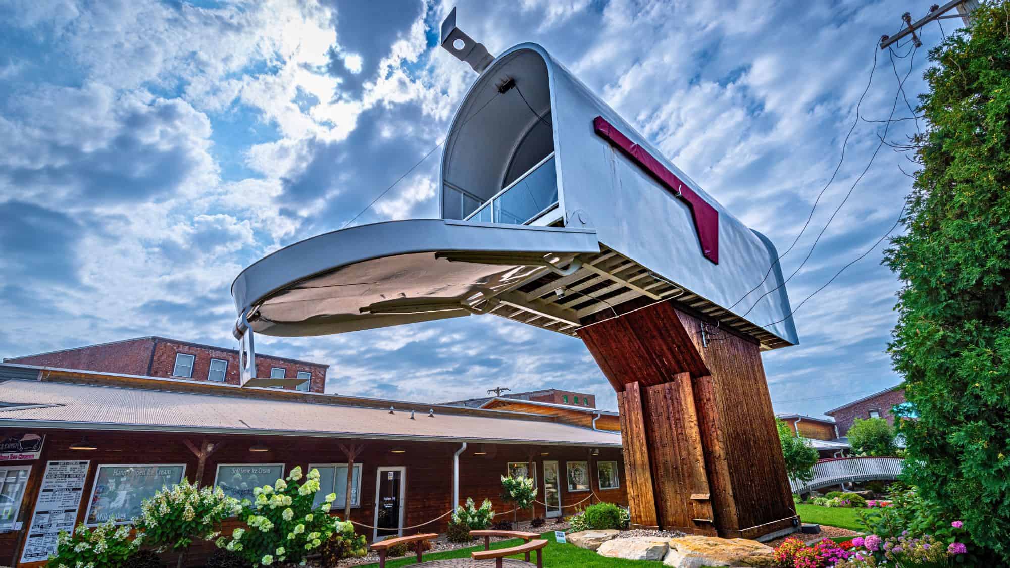 A towering, fully functional silver mailbox with a red flag and wooden base stands in front of a rustic downtown building, surrounded by flowers and benches.