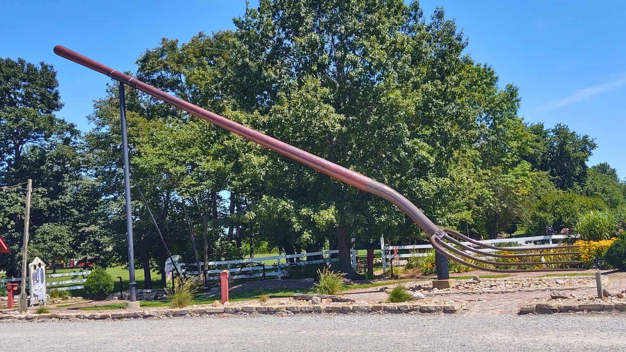 A colossal metal pitchfork sculpture leans diagonally on a black metal support, situated in a gravel and garden area with a white fence and trees in the background.