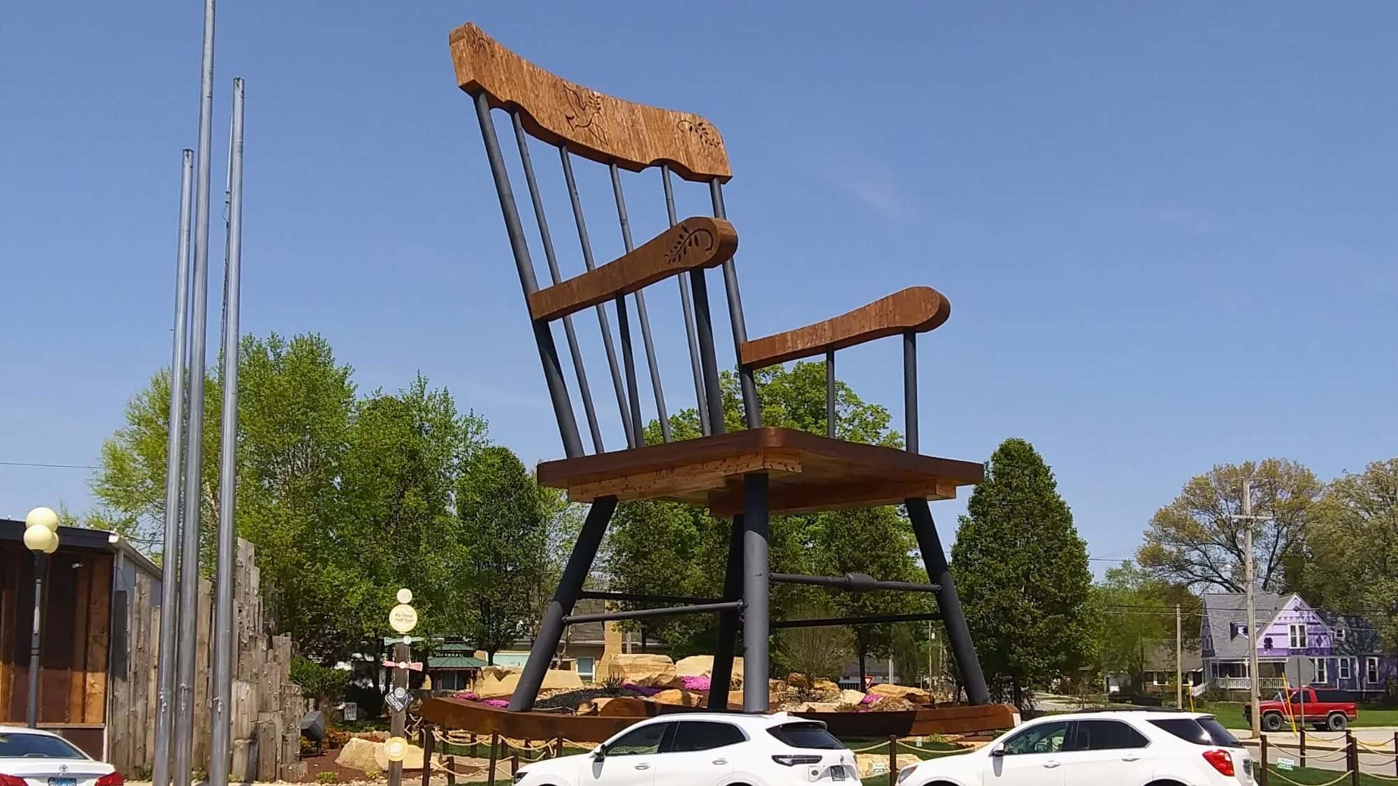 An enormous wooden and metal rocking chair sits elevated on a landscaped platform in the middle of a small town, with parked cars below for scale.