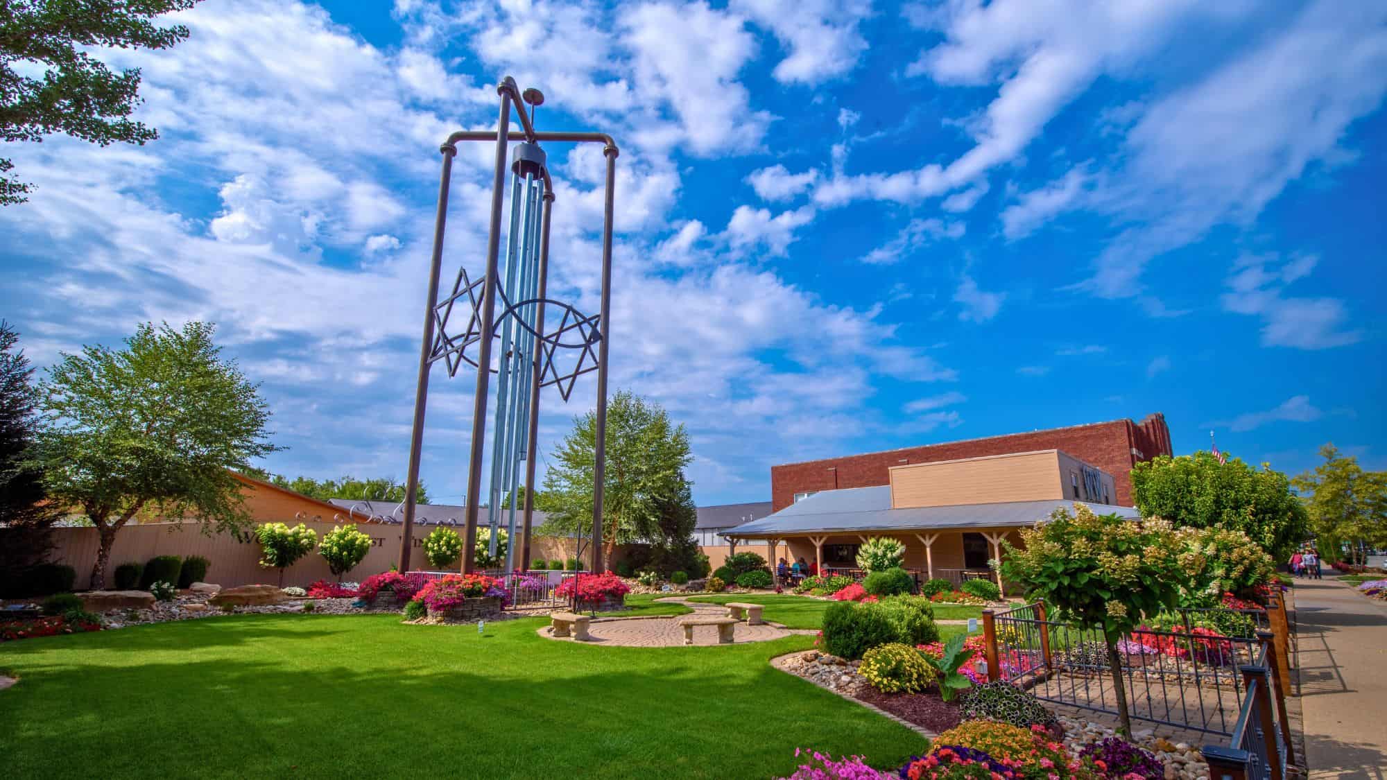 A towering set of wind chimes made of long silver pipes stands in a lush garden with colorful flowers, a stone pathway, and a blue sky above.