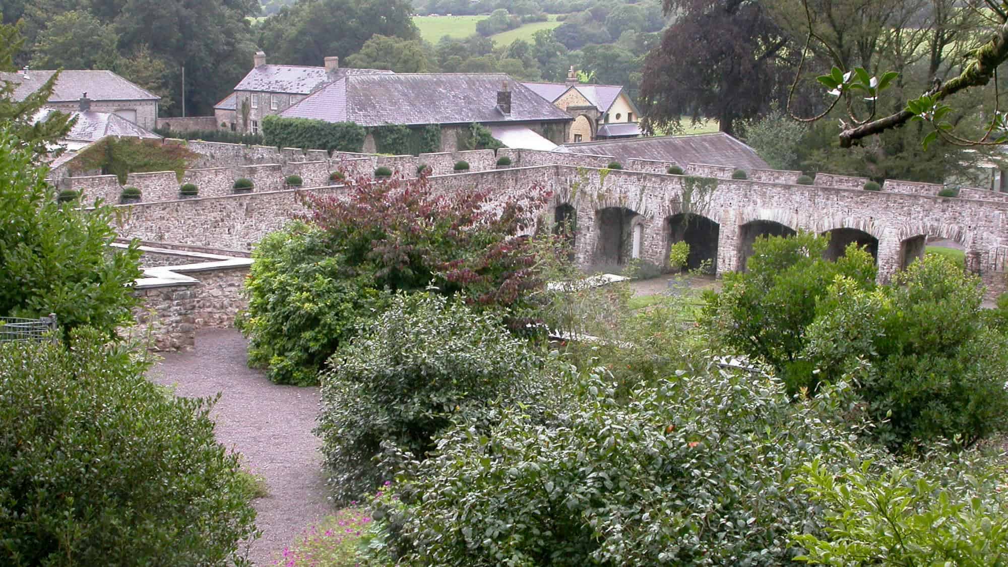 View of Aberglasney's beautifully preserved stone garden walls with archways and landscaped greenery, backed by charming Welsh stone buildings and lush countryside.
