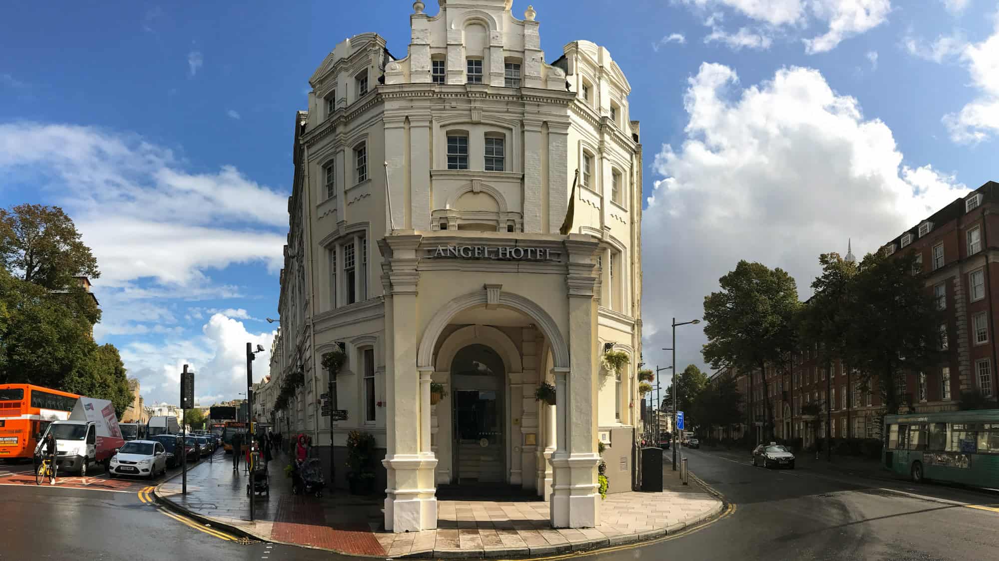 A grand corner hotel with white-painted stonework and arched entryway reading “Angel Hotel,” situated at a busy city intersection under a bright, partly cloudy sky in Cardiff, Wales.
