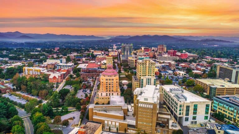 An aerial view of downtown Asheville, North Carolina at sunset, with the Blue Ridge Mountains in the background and a colorful skyline reflecting the day’s end.