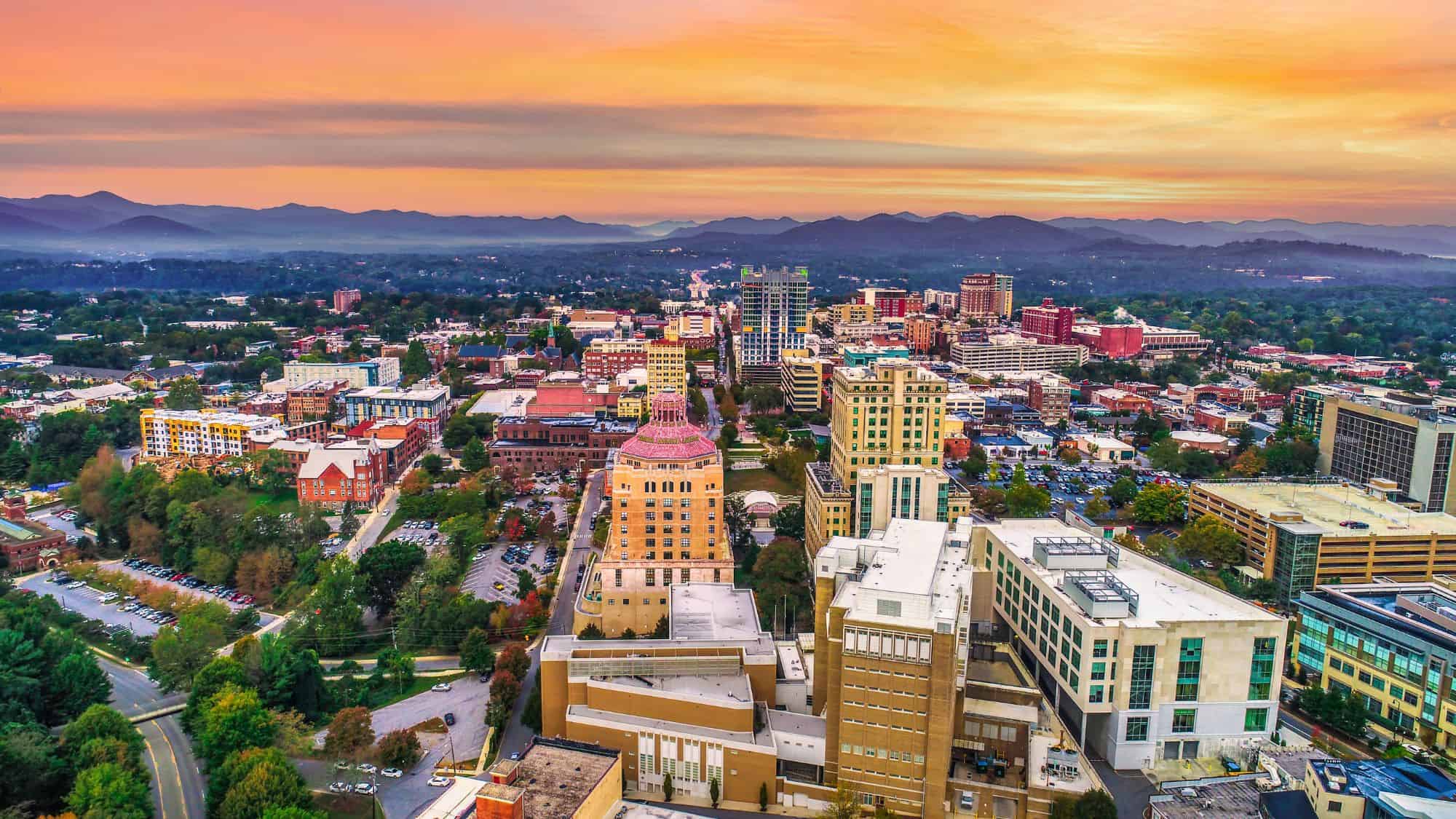 An aerial view of downtown Asheville, North Carolina at sunset, with the Blue Ridge Mountains in the background and a colorful skyline reflecting the day’s end.