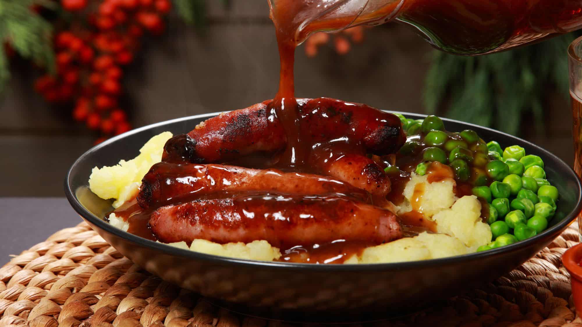 Grilled sausages smothered in rich brown gravy, served with mashed potatoes and green peas, with the gravy being poured over the dish.