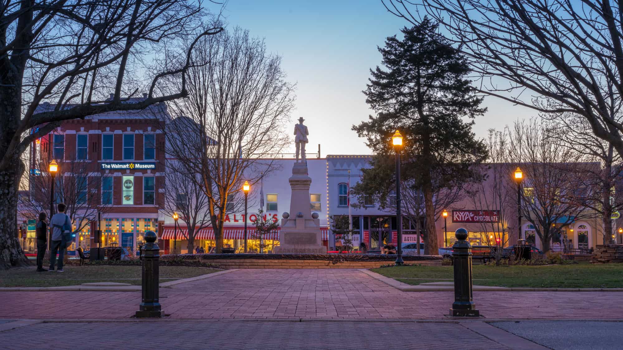 A twilight scene of Bentonville’s town square, featuring "The Walmart Museum," illuminated storefronts, bare trees, and a central statue of a soldier against a gradient blue and orange sky.