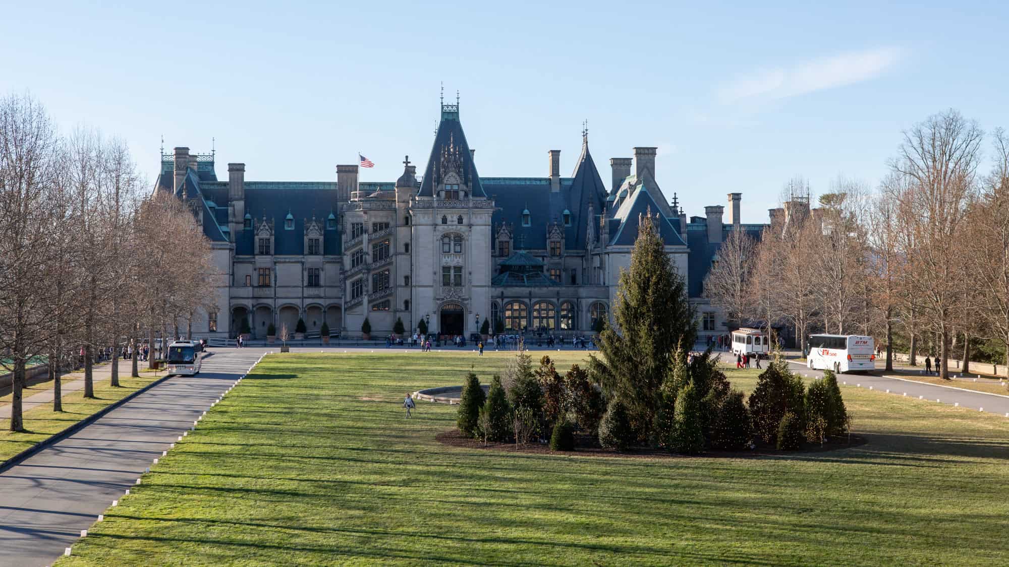 The grand Biltmore House in Asheville, North Carolina stands in the distance, framed by a sweeping driveway, trimmed lawns, and bare winter trees under a clear sky.