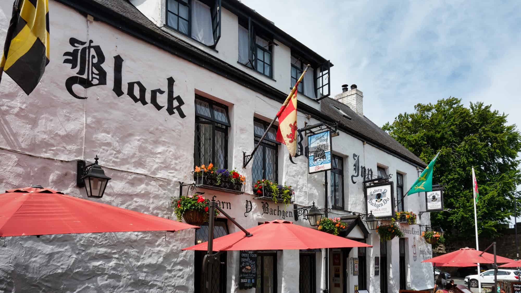 A traditional whitewashed inn with black medieval lettering and flags flying above a charming patio of red umbrellas and flower boxes. The sign reads “Black Boy Inn – Circa 1522,” showcasing its long-standing Welsh hospitality.