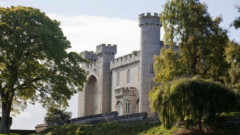 View of Bodelwyddan Castle’s grey stone facade with battlemented towers, partly obscured by mature trees, sitting atop a slight hill bathed in soft afternoon light.