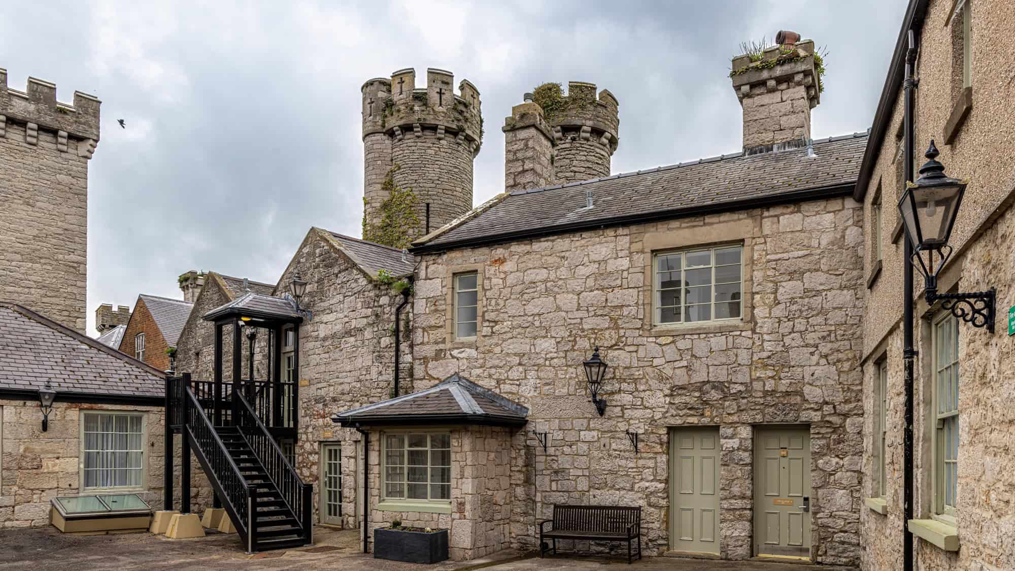 A quiet stone courtyard surrounded by medieval-style architecture and round battlement towers, with black stairs leading to an upper floor and classic lampposts enhancing the castle charm.