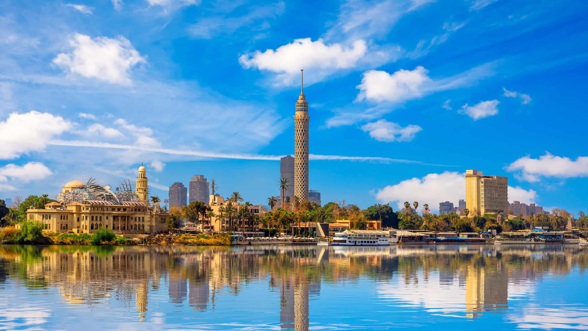 A view of Cairo, Egypt, featuring the iconic Cairo Tower and a mix of historic and modern architecture. The Nile River in the foreground reflects the cityscape against a bright blue sky.