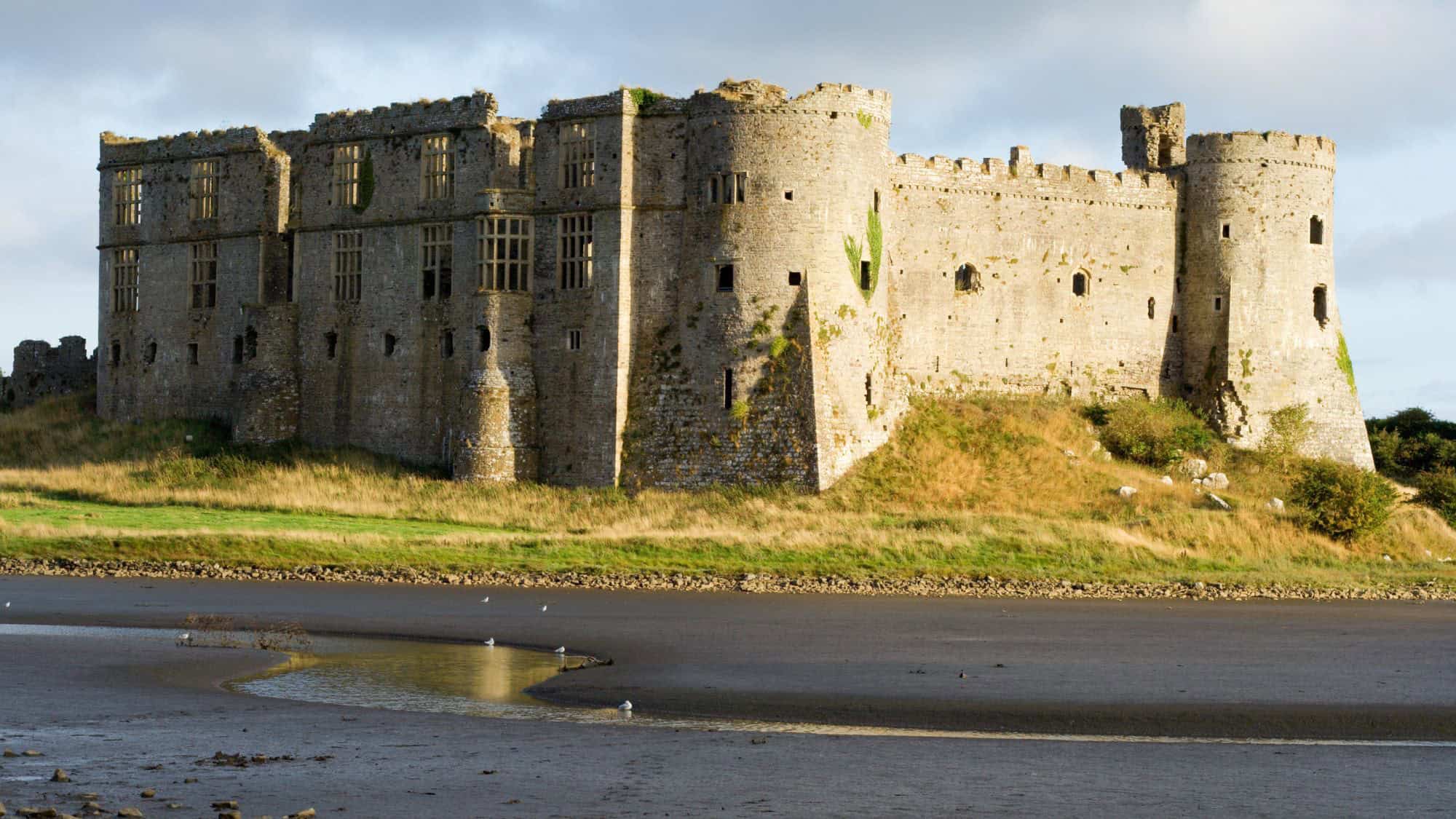 A weathered stone fortress sits on a grassy hill beside a tidal inlet, its medieval towers and narrow windows standing strong above the moody, low-tide landscape.