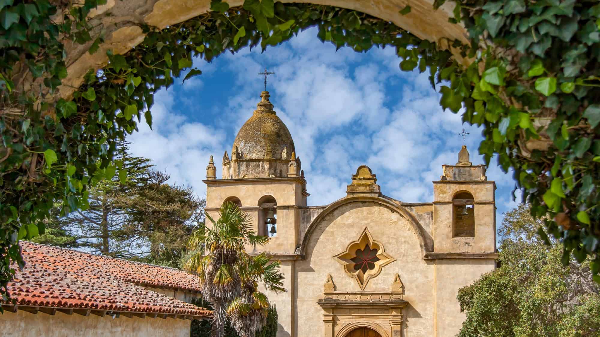 The ornate façade and bell towers of the Carmel Mission Basilica are framed by ivy-covered stone, with a clear blue sky and a hint of Spanish colonial charm.