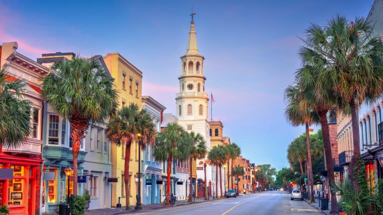 A charming street scene in Charleston, South Carolina, featuring pastel-colored historic buildings, palm trees, and a church steeple set against a soft sunset sky.