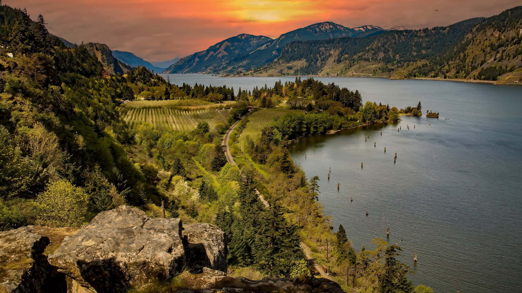 A breathtaking view of the Columbia River Gorge at sunset, showcasing lush green hills, farmland, and a winding river with distant mountains silhouetted against a golden sky.