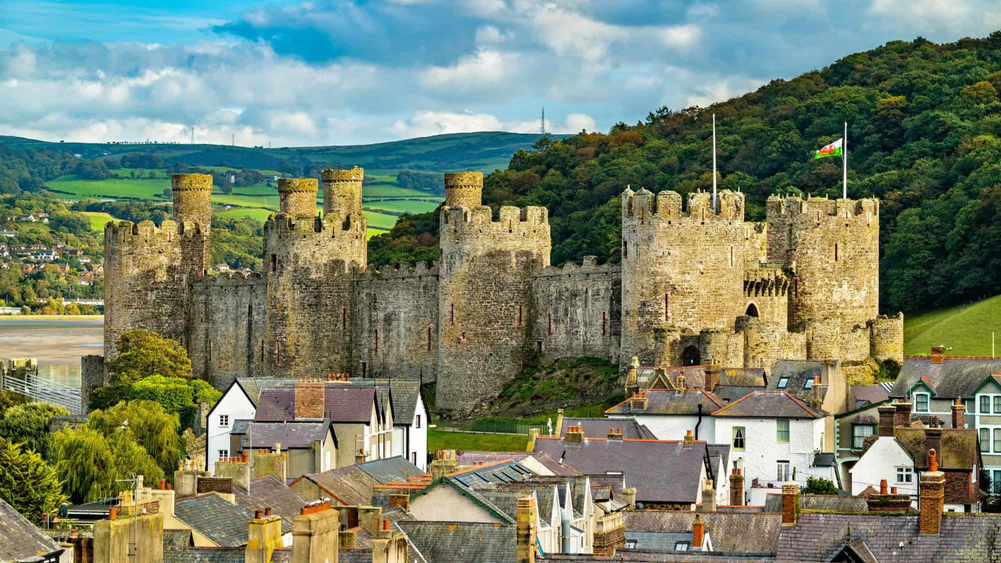 The majestic Conwy Castle rises above rooftops and nestled against forested hills, with its round stone towers, battlements, and flying Welsh flag standing guard over the nearby estuary.