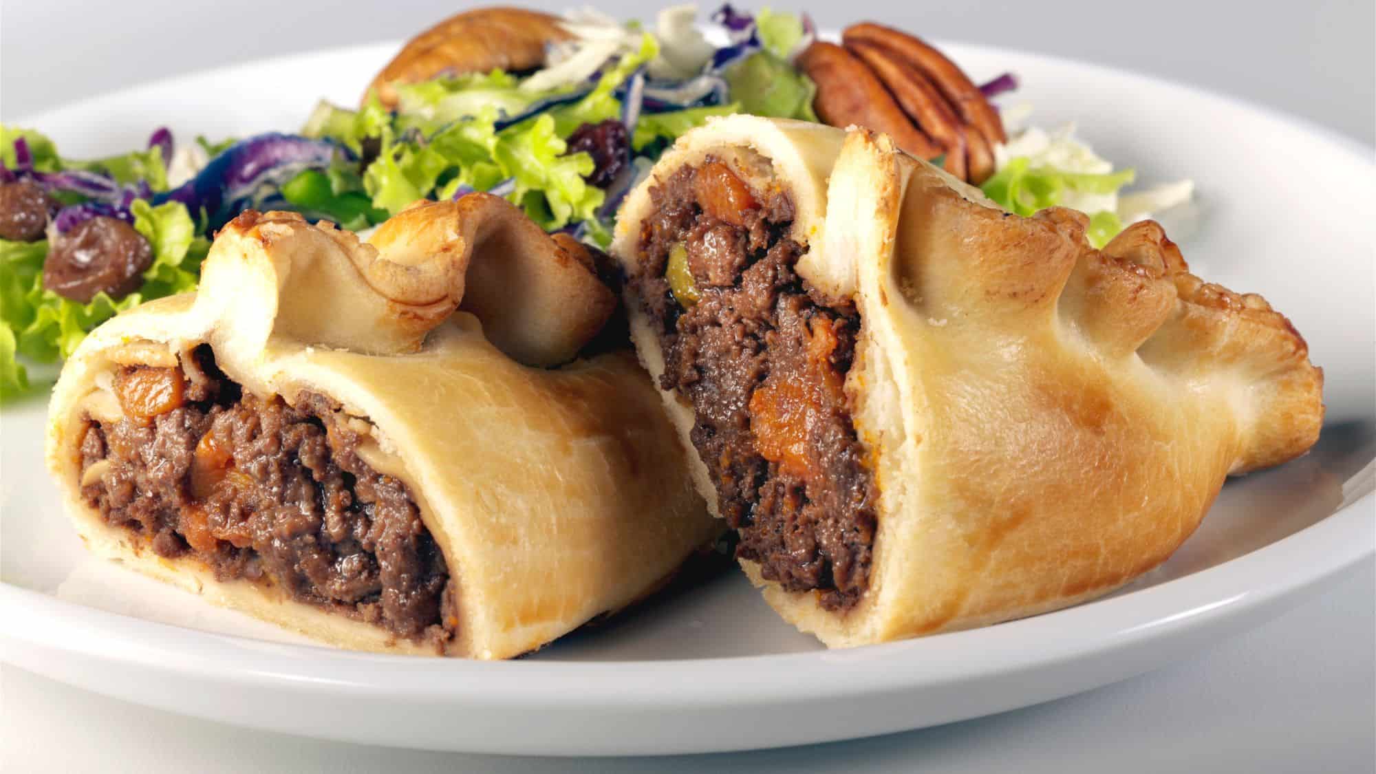 A flaky, golden-brown pastry filled with minced beef, potatoes, carrots, and onions, cut in half to reveal the rich, savory filling, accompanied by a fresh green salad.