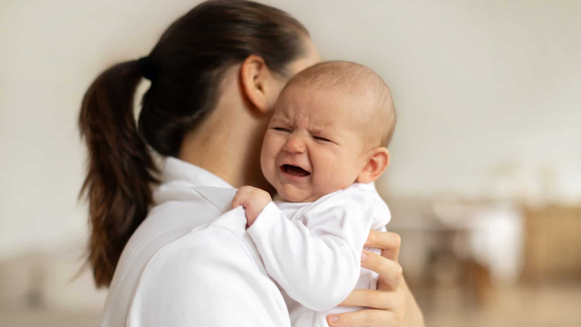 A mother dressed in white holds her crying baby close to her shoulder. The baby, also in white, has a distressed expression with furrowed brows and an open mouth.