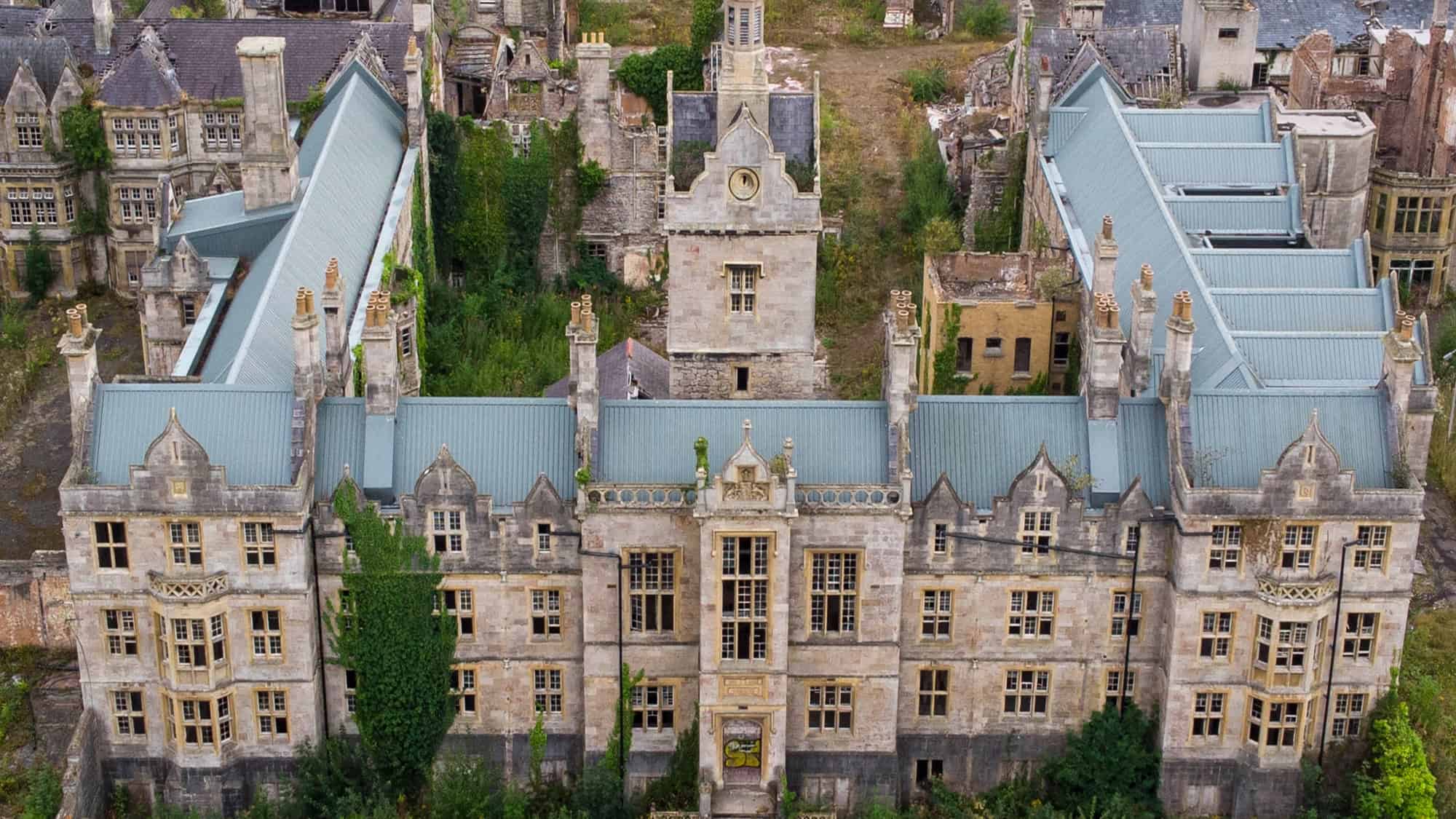 A haunting view of the abandoned Denbigh Asylum, with decaying Gothic Revival buildings overtaken by ivy and nature, its tall chimneys and stonework hinting at its grand but eerie past.