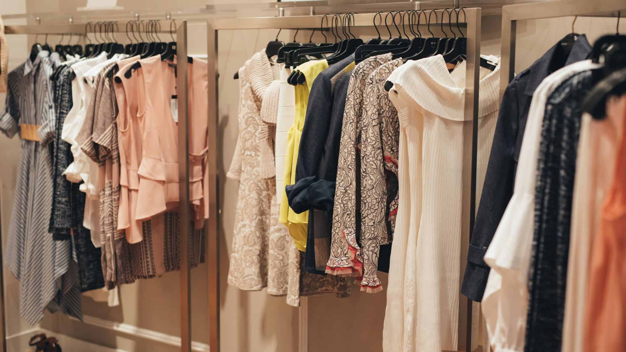A neatly arranged clothing rack displays an assortment of elegant women's garments, including lace dresses, pastel blouses, and knitwear, in a stylish boutique.

