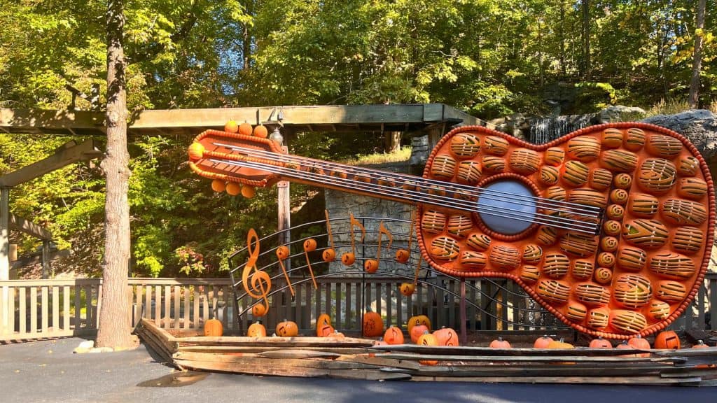 A massive guitar sculpture made of intricately carved pumpkins stands against a lush green forest backdrop. The display is accented with metal musical notes and a wooden fence, celebrating a festive autumn theme.