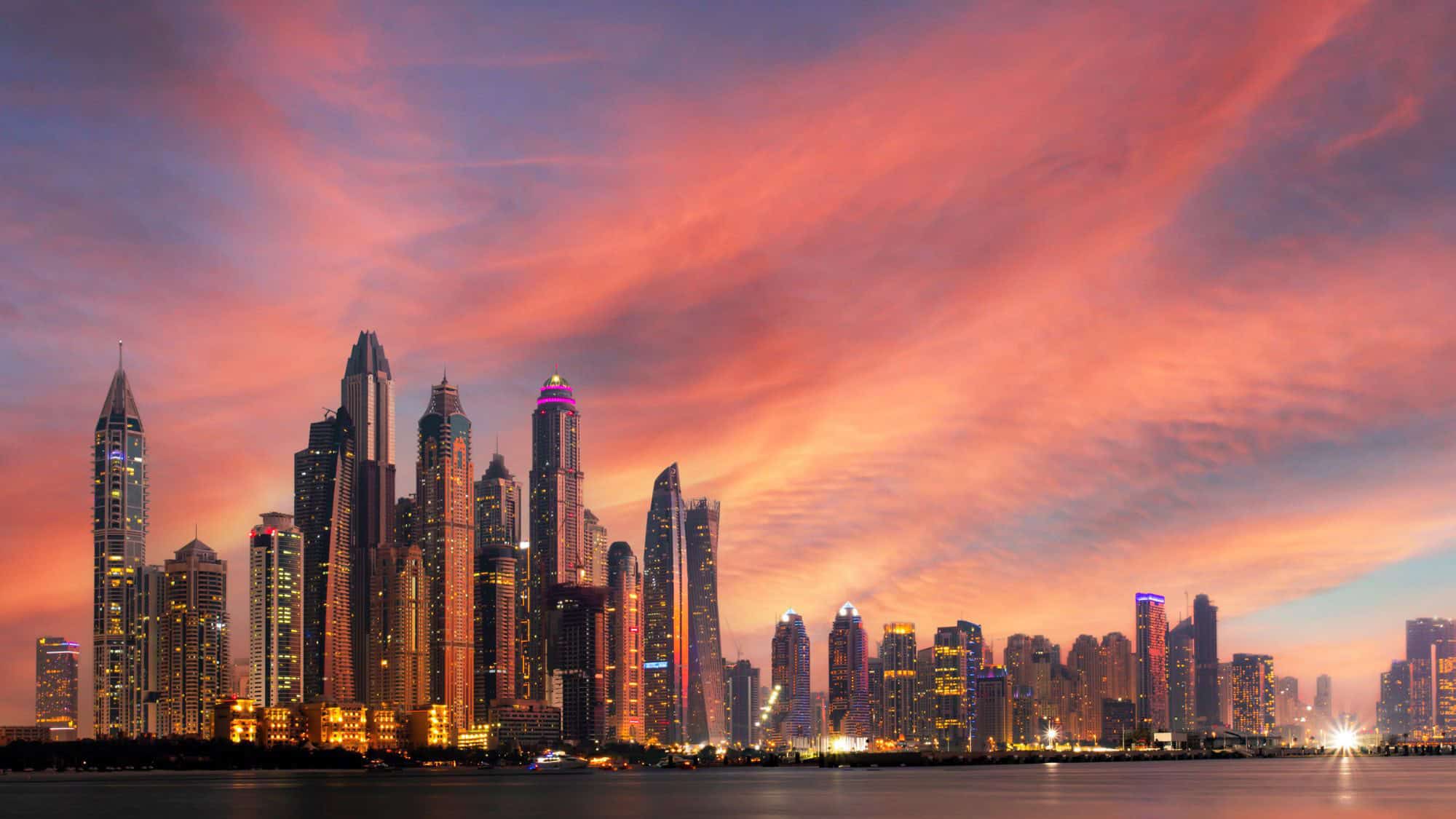 A breathtaking view of Dubai’s skyline at dusk, with the city's towering skyscrapers illuminated against a vivid pink and orange sunset sky. The buildings reflect on the calm waters in the foreground.