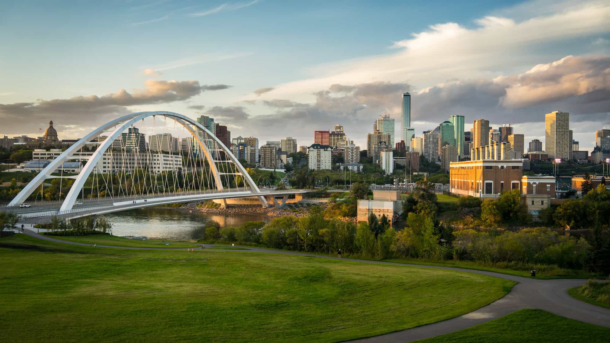 A picturesque view of Edmonton’s skyline, with the Walterdale Bridge stretching across the North Saskatchewan River, modern high-rises, and lush greenery in the foreground under a partly cloudy sky.