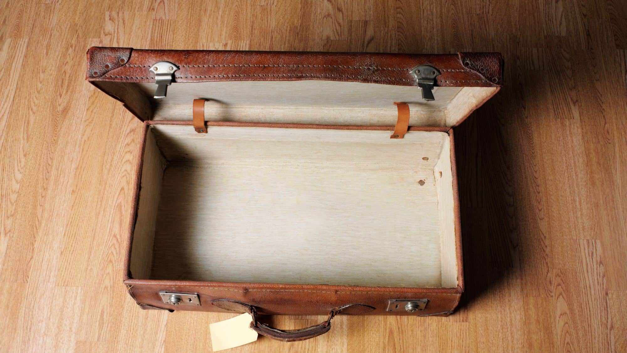 A vintage brown leather suitcase sits open on a wooden floor, revealing its empty, cream-colored interior with leather straps.
