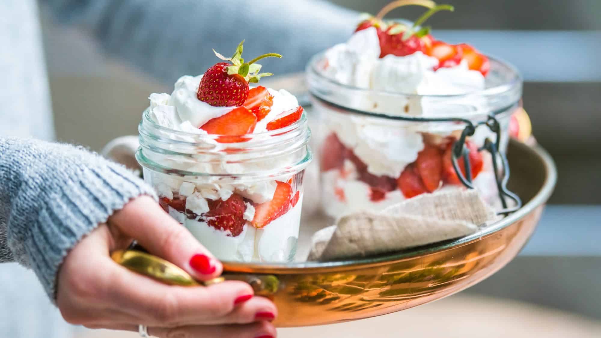 A refreshing British dessert consisting of crushed meringue, whipped cream, and fresh strawberries, served in mason jars on a brass tray.