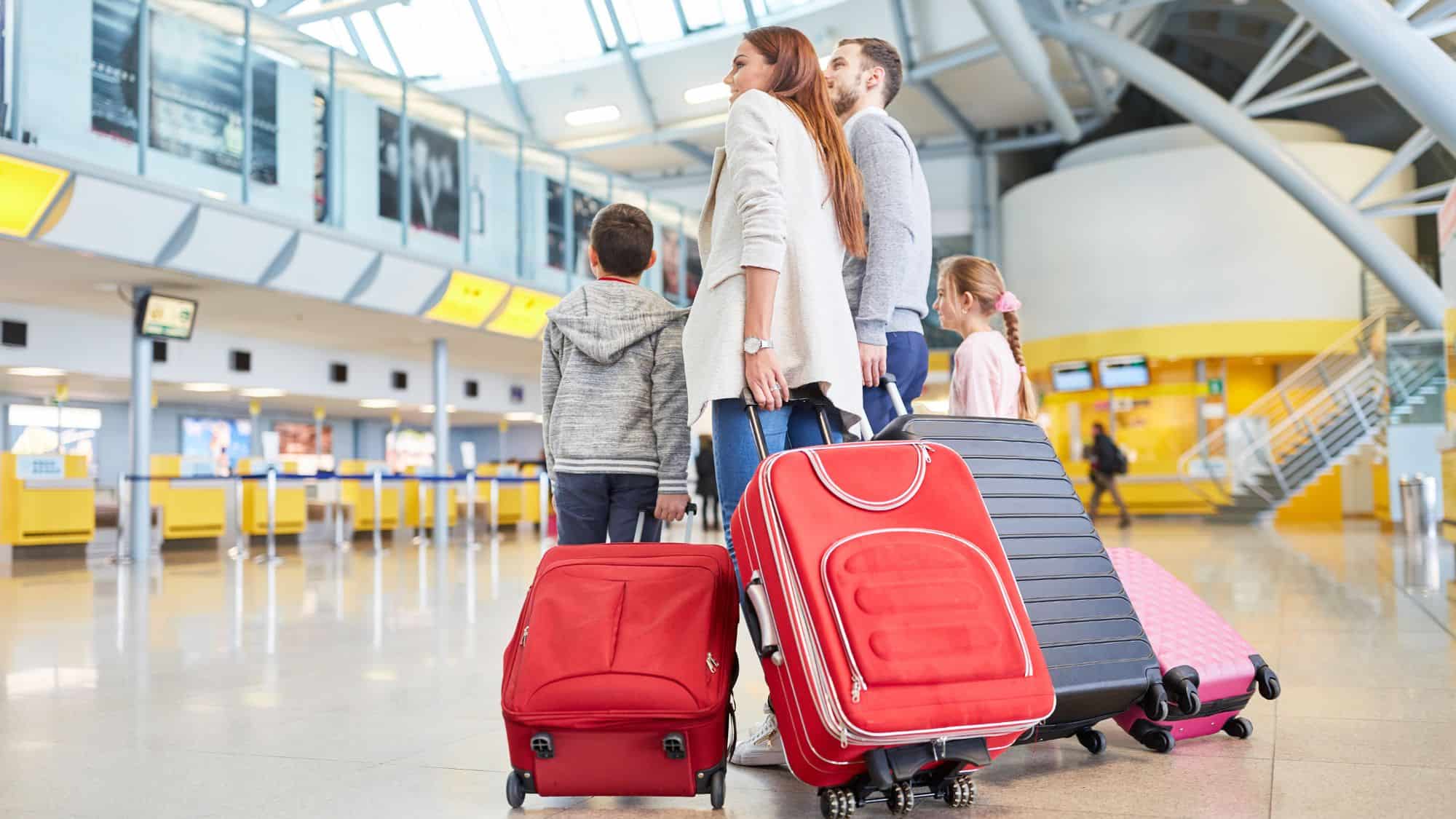 A family of four walks through a bright and modern airport terminal, pulling rolling suitcases in red, black, and pink. The parents and children look ahead toward the check-in counters.