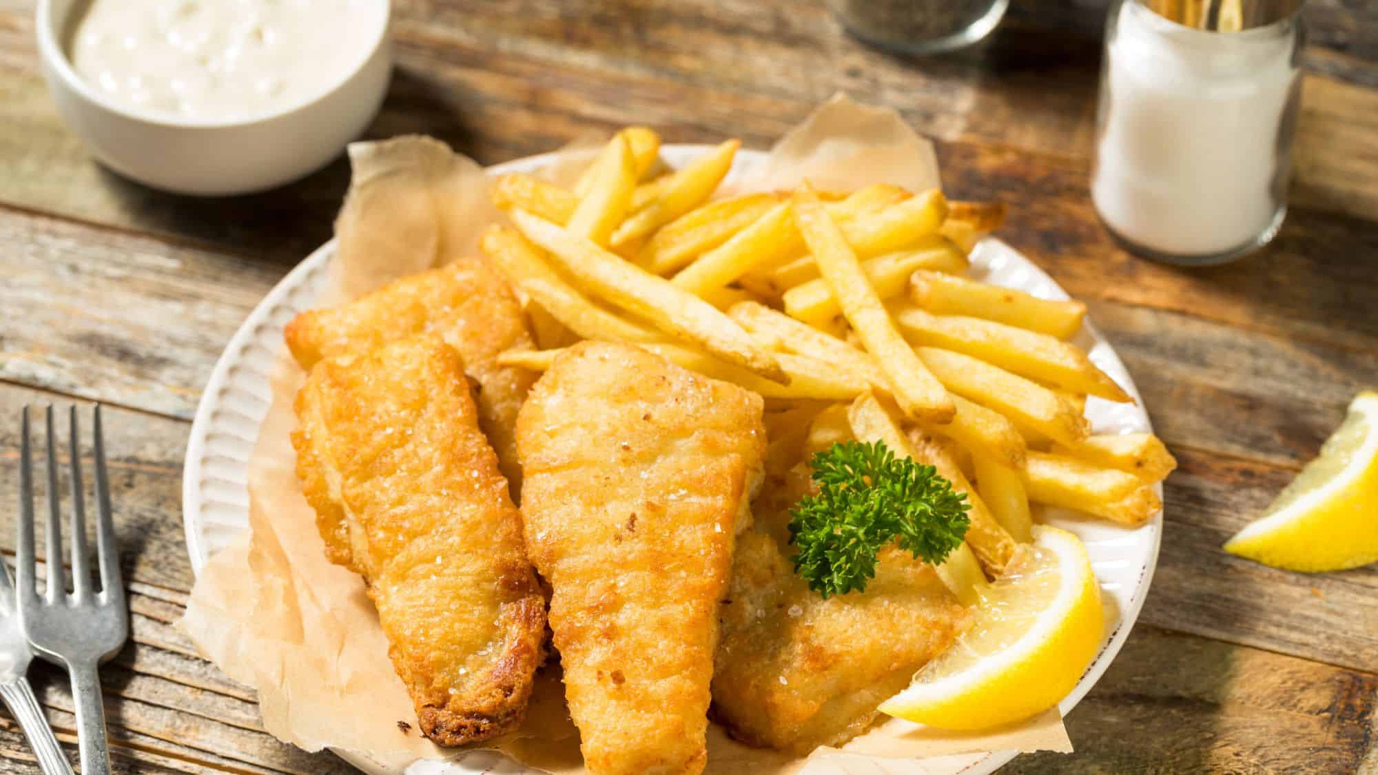 A classic British dish with golden, crispy battered fish fillets served alongside thick-cut fries, lemon wedges, and a side of tartar sauce on a rustic wooden table.