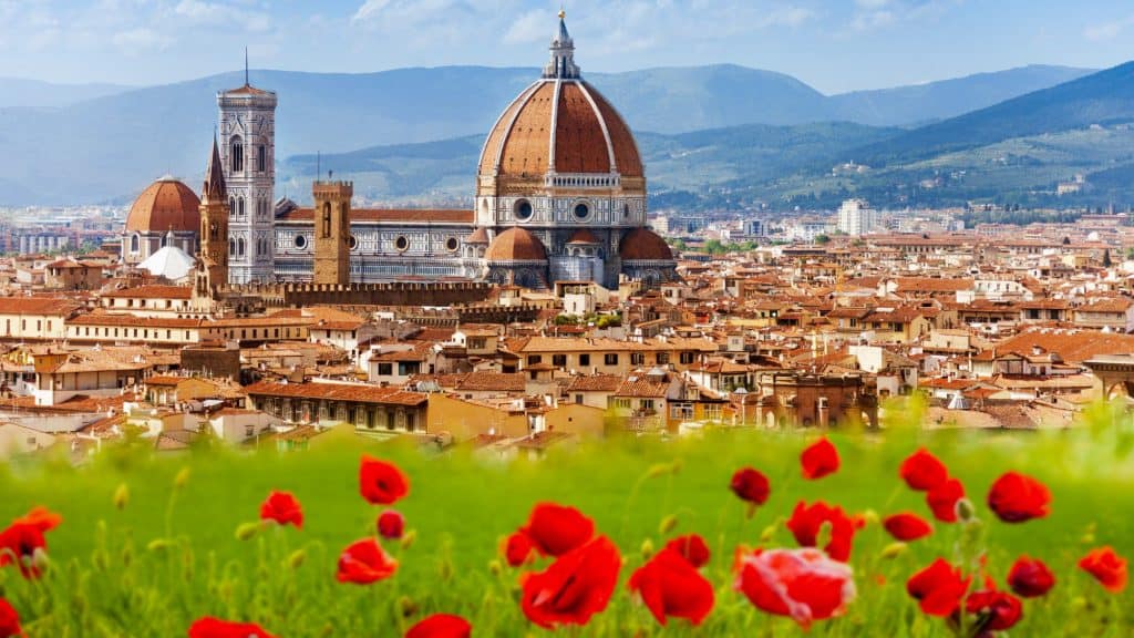 A picturesque scene of Florence, Italy, featuring the iconic red-domed Cathedral of Santa Maria del Fiore. In the foreground, a lush green field is dotted with vibrant red poppies, adding contrast to the historic skyline.