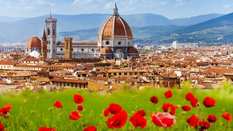 A picturesque scene of Florence, Italy, featuring the iconic red-domed Cathedral of Santa Maria del Fiore. In the foreground, a lush green field is dotted with vibrant red poppies, adding contrast to the historic skyline.