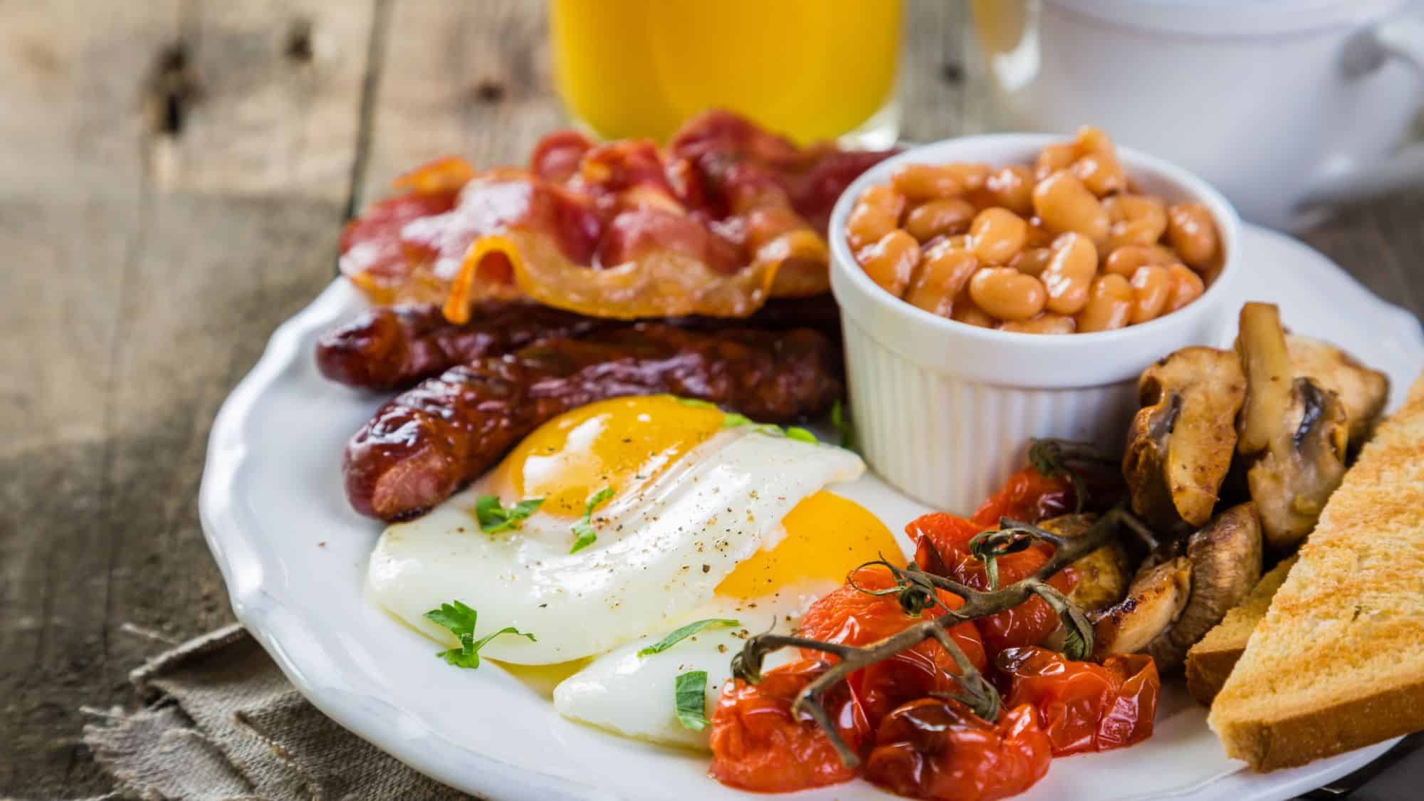 A hearty plate featuring crispy bacon, sausages, sunny-side-up eggs, baked beans, grilled tomatoes, sautéed mushrooms, and buttered toast, served with a cup of tea or orange juice.