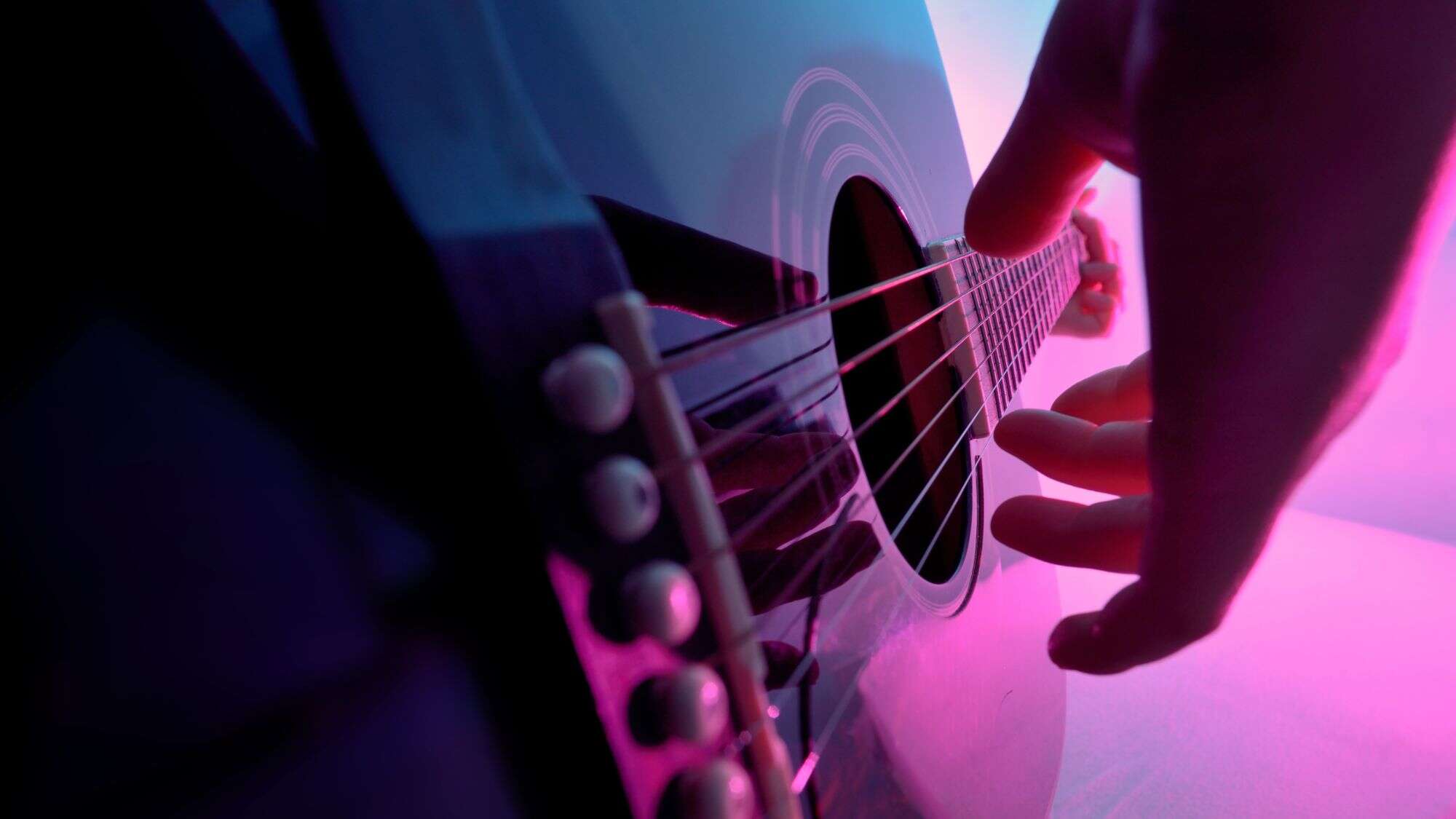 A close-up of a musician's hands strumming an acoustic guitar, illuminated by moody pink and purple lighting. The perspective focuses on the strings and fingers, emphasizing the texture and motion.