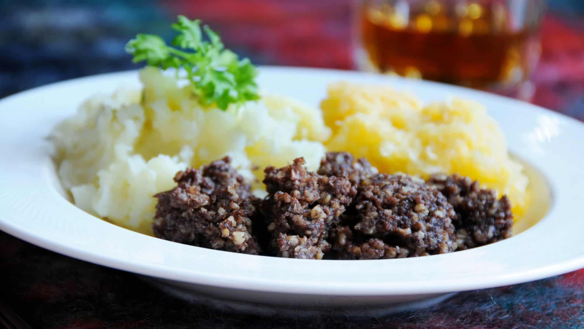 A traditional Scottish dish featuring haggis served with mashed potatoes and turnips, garnished with fresh parsley, and accompanied by a glass of whisky in the background.