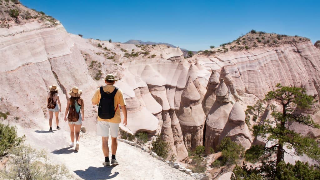 A group of hikers wearing sun hats and backpacks explore a scenic trail at Kasha-Katuwe Tent Rocks National Monument. The towering rock formations and rugged desert landscape surround them, highlighting the area's geological beauty.
