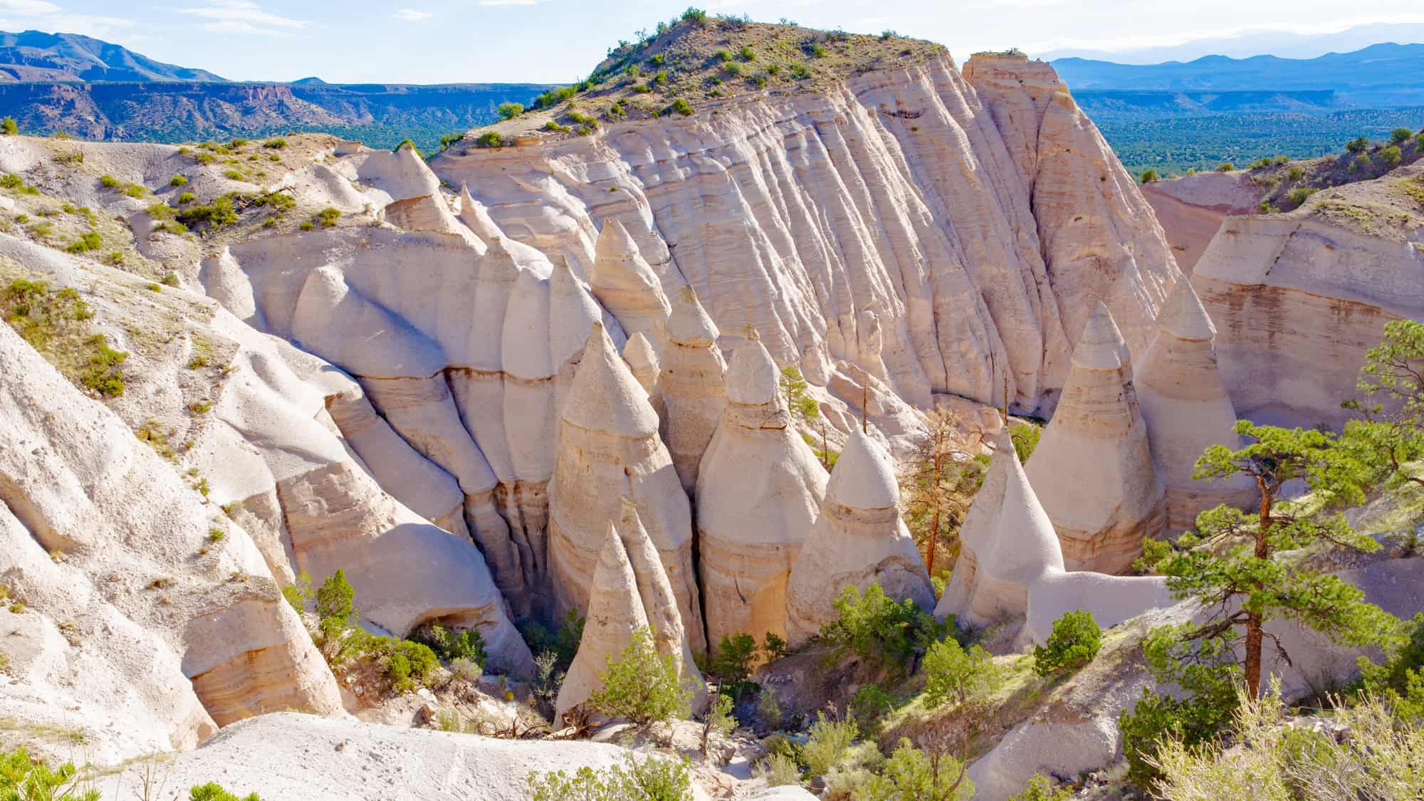 A stunning landscape of cone-shaped rock formations at Kasha-Katuwe Tent Rocks National Monument, New Mexico. The light-colored cliffs and towering hoodoos stand against a blue sky, surrounded by sparse vegetation.